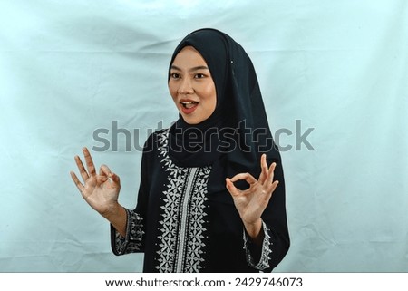 beautiful young Asian Muslim woman wearing hijab and black dress with white pattern showing okay sign. smile showing her teeth isolated on white background