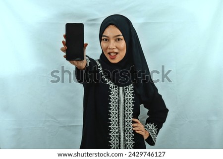 beautiful young Asian Muslim woman wearing hijab and black dress with white pattern showing blank screen smartphone isolated on white background
