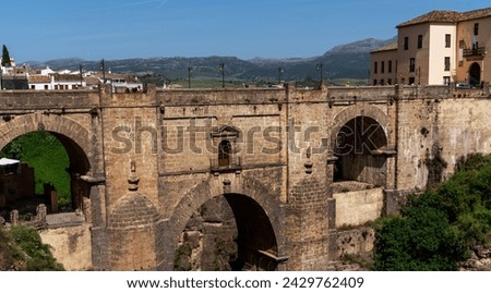 An ancient stone bridge arches majestically over a chasm, connecting two parts of an old town Ronda against a backdrop of rolling hills and clear skies. Royalty-Free Stock Photo #2429762409