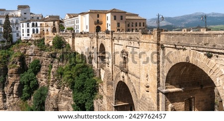 An ancient stone bridge arches majestically over a chasm, connecting two parts of an old town Ronda against a backdrop of rolling hills and clear skies. Royalty-Free Stock Photo #2429762457