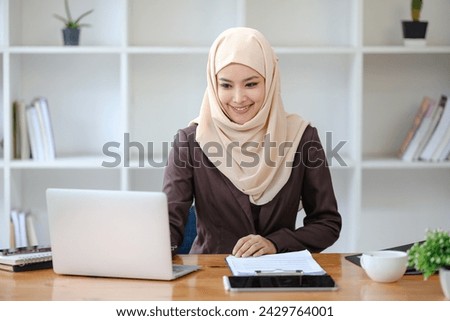 young pretty modern muslim woman in hijab working on laptop in office room, education online, remote work freelancer, using smartphone