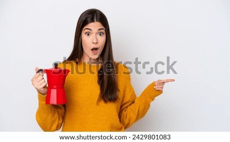 Young Brazilian woman holding coffee pot isolated on white background surprised and pointing side