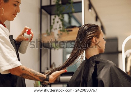 Beautiful hair stylist with tattooed hands applying spray on woman wet hair in beauty salon Royalty-Free Stock Photo #2429830767