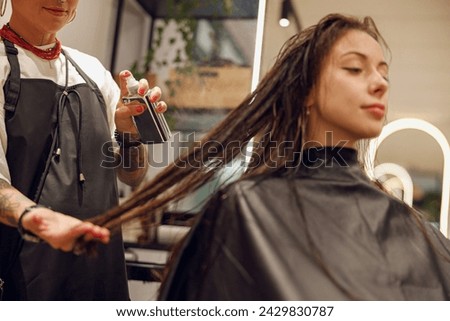 Beautiful hair stylist with tattooed hands applying spray on woman wet hair in beauty salon Royalty-Free Stock Photo #2429830787