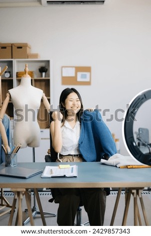 Beautiful Asian lady blogger showing clothes in front of the camera to recording vlog video live streaming at her shop. Online Shopping Cart notion.
