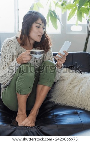 A relaxed woman enjoys her coffee while browsing on her phone, a picture of modern leisure. Wrapped in a warm knit sweater and comfortably seated, she embodies the simplicity of a peaceful morning in 