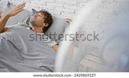 Hispanic man with beard enjoys breeze from fan while lying in bedroom, eyes closed in relaxation Royalty-Free Stock Photo #2429869237