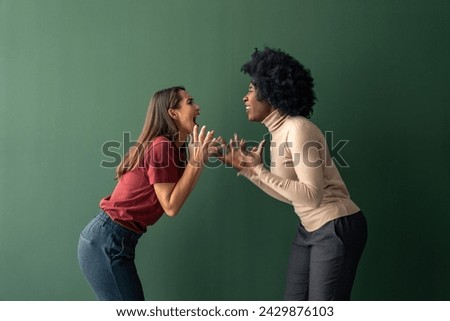 Two young interracial women yelling at each other against a green background in a studio.