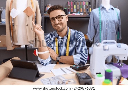 Hispanic man with beard dressmaker designer working at atelier with a big smile on face, pointing with hand finger to the side looking at the camera. 