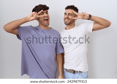Homosexual gay couple standing over white background doing peace symbol with fingers over face, smiling cheerful showing victory 