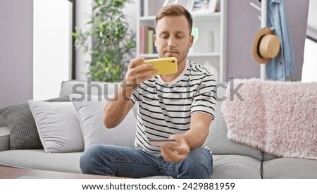 Relaxed yet serious young caucasian man, sitting on living room sofa, taking photo of credit card with phone, a smart step into online shopping realm at home