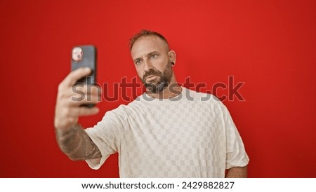 Cool, casual young guy making a serious-face selfie over a vivid, isolated red background with his slick smartphone
