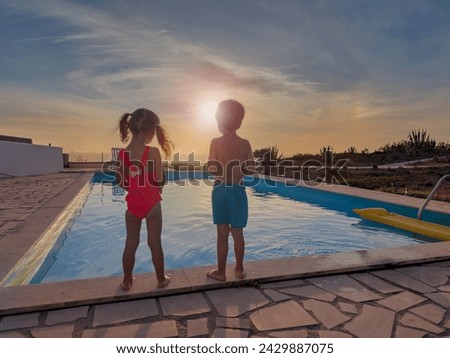 Two children stand at the edge of a swimming pool, looking at a sunset their figures outlined by the retreating sunlight