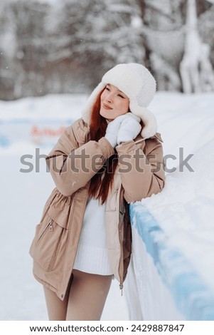 young pretty caucasian redhead smiling girl in white and beige winter clothes skating in ice rink in winter cloudy snowy day, standing and resting