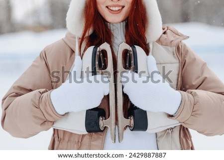 young pretty caucasian redhead girl in white and beige winter clothes skating in ice rink in winter cloudy snowy day, skates in hands, smiling widely, close-up view