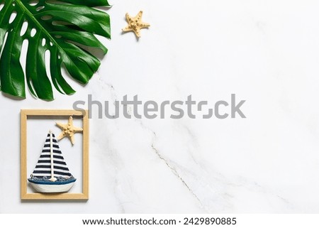 Summer vacation concept. Artistic composition with big tropical monstera leaf, decorative star fish and  wooden picture frame with a boat on white marble background. Copy space for your design.