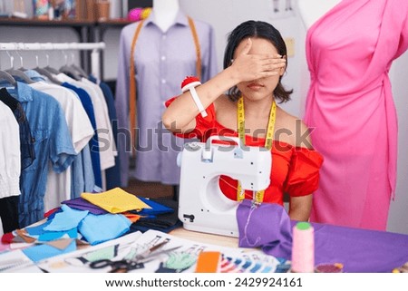 Hispanic young woman dressmaker designer using sewing machine covering eyes with hand, looking serious and sad. sightless, hiding and rejection concept  Royalty-Free Stock Photo #2429924161