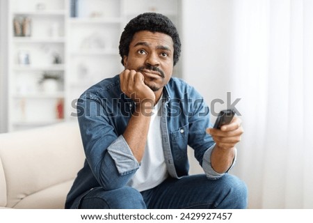 Portrait Of Bored Young Black Man Watching TV At Home, Discontented African American Guy With Remote Controller In Hand Sitting On Couch At Home, Leaning Head On Hand And Switching Channels