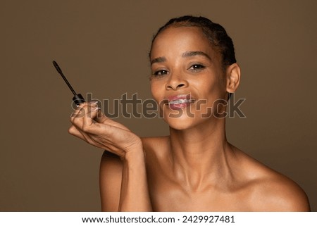 Cheerful african american middle aged woman with radiant smile holding mascara wand, ready to apply makeup in beauty routine, set against brown background