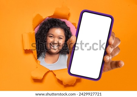 Cheerful african american plus size young woman with curly hair peering through a torn orange paper, presenting a smartphone with a blank screen, perfect for mockup designs