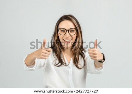 A smiling european young woman with long hair and glasses gives two enthusiastic thumbs up, wearing a white blouse and smartwatch, showing approval or success, studio, close up