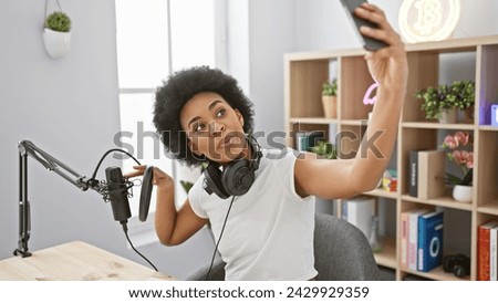 A cheerful african american woman takes a selfie in a modern radio studio setup with professional recording equipment.