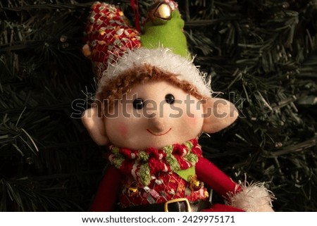 Close-up of a dressed-up elf plush Christmas ornament hanging from a tree with lights behind
