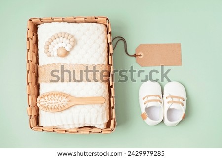 Top view of a wicker basket containing soft knitted blankets and natural wooden baby grooming tools, on a gentle green background with gift tag mockup