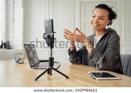 A joyful young woman creates content, gesturing to a smartphone on a tripod, with a laptop open in front of her.