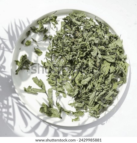 Sunlit dried mint leaves spread on a white plate, casting playful shadows. The herbs offer a promise of refreshing flavor and natural goodness.