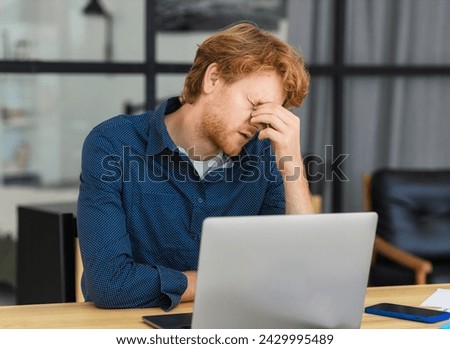 After working for an extended period on his laptop, the overtired businessman experiences a headache while sitting at his desk in the office Royalty-Free Stock Photo #2429995489