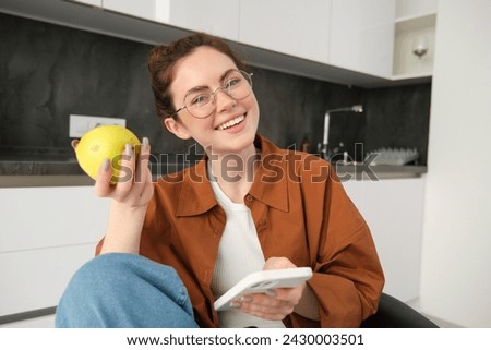 Image of young modern woman, relaxing at home, sitting on chair and using smartphone, eating green apple and smiling.