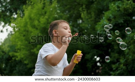 Preschooler boy spends time blowing soap bubbles creating playful atmosphere slow motion. Playing in summer city park cheerful boy happily blows bubbles. Young boy delights in blowing soap bubbles Royalty-Free Stock Photo #2430008603