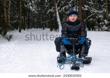 A boy on a sled looks directly into the camera against a forest backdrop. A child on a skibob against the background of the forest