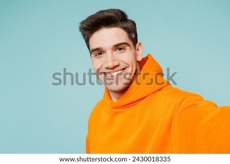 Close up smiling young man he wears orange hoody casual clothes doing selfie shot pov on mobile cell phone isolated on plain pastel light blue cyan color background studio portrait. Lifestyle concept