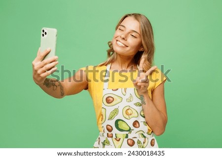 Young housewife housekeeper chef cook baker woman wear apron yellow t-shirt doing selfie shot on mobile cell phone show v-sign isolated on plain pastel green background studio. Cooking food concept