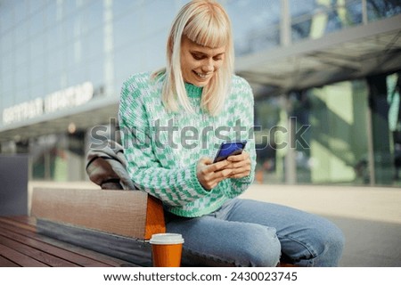 Portrait of a young female influencer sitting on a city street and typing on her phone. An urban girl is sitting on a bench downtown and using her cellphone for social media while smiling at the phone Royalty-Free Stock Photo #2430023745