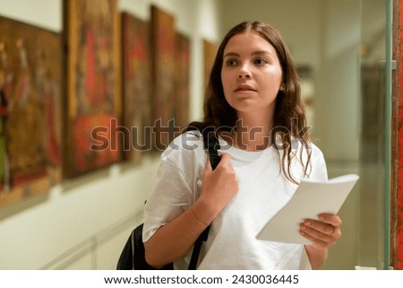 Portrait of a confident girl with an information booklet, standing in the hall of the historical museum next to the ..exhibit, located in a glass cabinet Royalty-Free Stock Photo #2430036445
