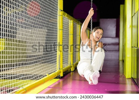 Young girl in white sportswear having fun in modern indoor trampoline amusement center, riding zip line against vibrant colorful background..