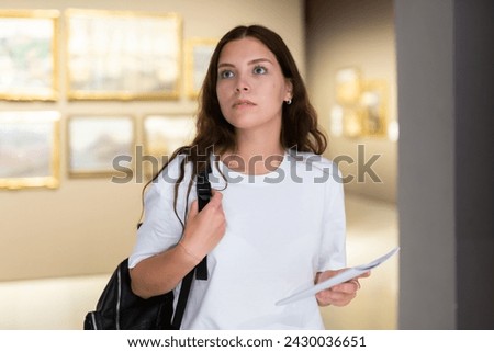 Portrait of a young girl visitor with a paper guide at the exhibition of paintings in the museum Royalty-Free Stock Photo #2430036651