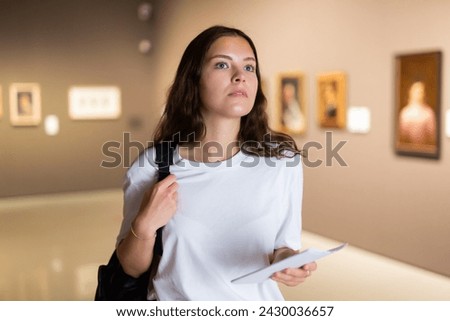 Thoughtful young girl visiting an art gallery exhibition of paintings studies the artwork with information booklet..in her hand Royalty-Free Stock Photo #2430036657