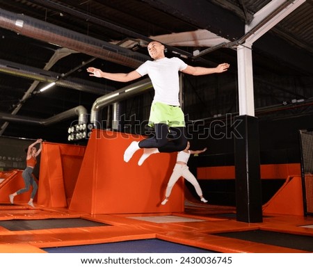 Energetic young man exuding joy while leaping at trampoline park, showcasing carefree fun of indoor recreational activity.. Royalty-Free Stock Photo #2430036745