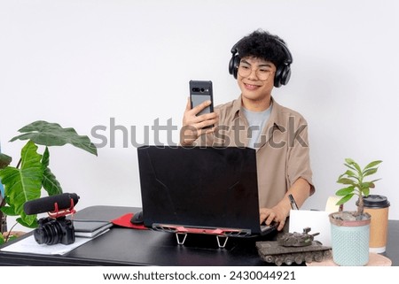 A cheerful young Asian freelancer video editing at his desk, with headphones on and taking a break to use his smartphone. Taking a video call.