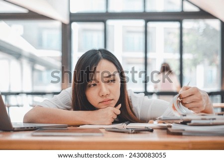 A weary woman rests her head on her hand, looking overwhelmed by work as she sits at a desk filled with papers and a laptop. Royalty-Free Stock Photo #2430083055