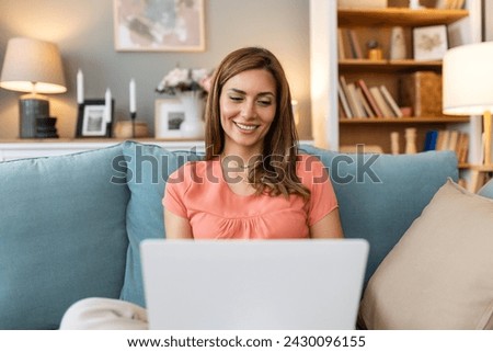 Young woman doing research work for her business. Smiling woman sitting on sofa relaxing while browsing online shopping website. Happy girl browsing through the internet during free time at home.