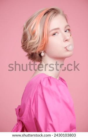 A cute blonde teenage girl with a short haircut poses in a pink dress and makes a bubble with gum. Pink background. Lovely spring-summer look.