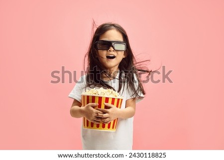 Joyful and Surprised Child Girl in 3D Glasses Holding Popcorn Bucket Having Flowing Hair Standing Over Pink Isolated Background.