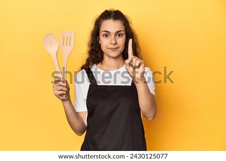 Woman with apron, wooden cooking utensils, yellow, showing number one with finger.