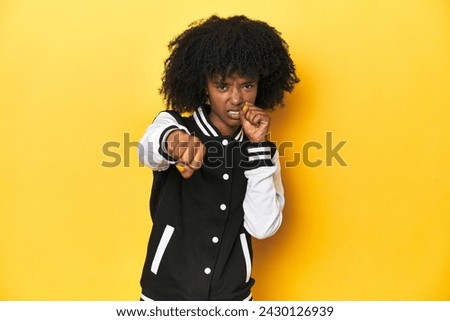 Teen girl in baseball jacket, yellow studio background throwing a punch, anger, fighting due to an argument, boxing. Royalty-Free Stock Photo #2430126939