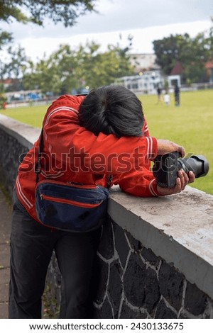 A man in a red jacket is taking photos using a camera in a city park.